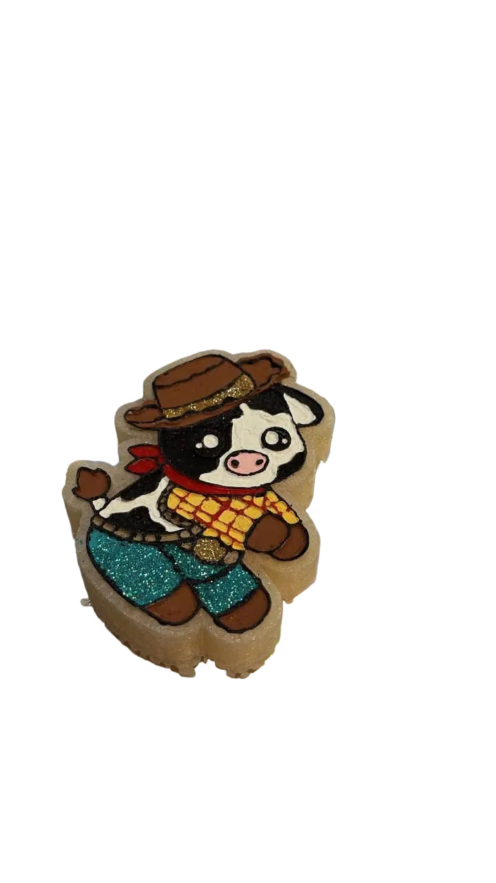 A small illustrated cow character accessory on a shiny holographic zip pouch, photographed on a wooden surface. The pouch has partial text reading 'Fragile Scents' and 'Beast'.